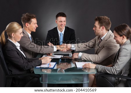 Businessmen shaking hands during conference meeting with colleagues at desk against black background