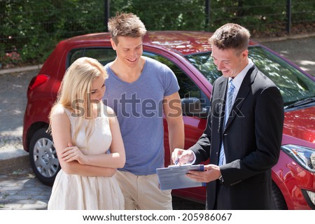 Young salesman showing contract paper to couple by new car