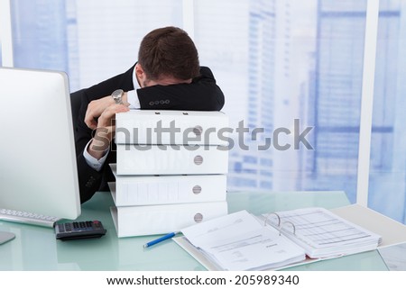 Stressed young businessman resting head on stacked binders at office desk