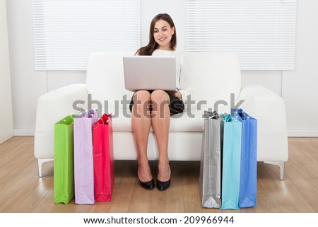 Full length of young businesswoman using laptop with shopping bags on floor at home