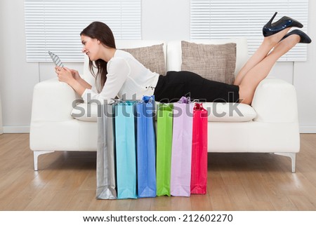 Full length side view of young businesswoman using digital tablet with shopping bags on floor at home