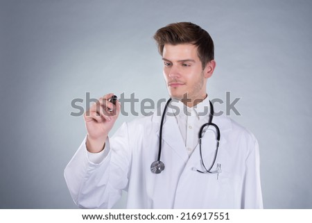 Young male doctor writing on transparent screen over gray background