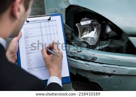 Side view of writing on clipboard while insurance agent examining car after accident