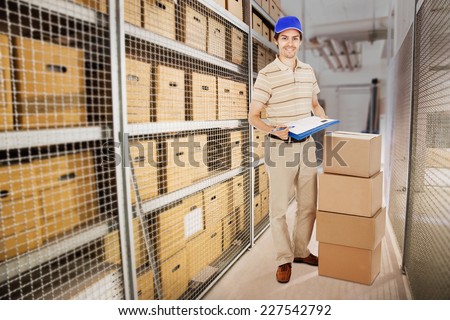 Full length portrait of smiling delivery man holding clipboard while standing by stacked cardboard boxes in warehouse