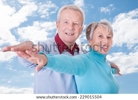 Portrait of happy senior couple with arms outstretched standing against cloudy sky