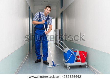 Full length portrait of happy male worker with broom cleaning office corridor