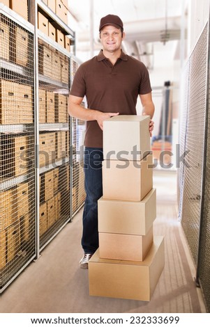 Portrait of smiling delivery man with stacked cardboard boxes standing in warehouse