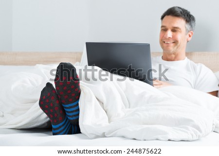 Happy Mature Man With Socks In His Leg Sitting On Bed Using Laptop