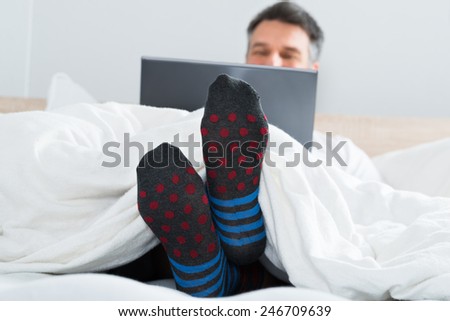 Mature Man With Socks In His Leg Sitting On Bed Using Laptop