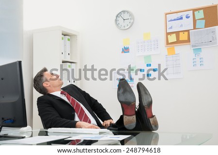 Mature Businessman With Feet On Desk Looking At Time In Office