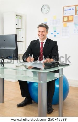 Businessman Writing On Paper Sitting On Pilates Ball In Office