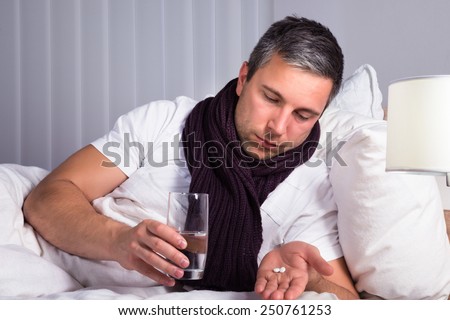 Sick Man On Bed Holding Glass Of Water And Looking At Pills