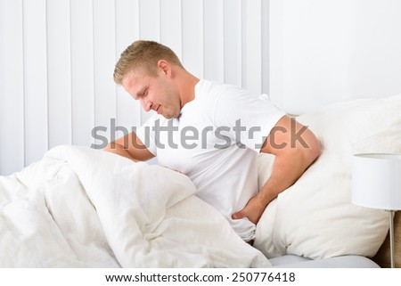 Portrait Of Young Man Sitting On Bed Suffering From Backpain