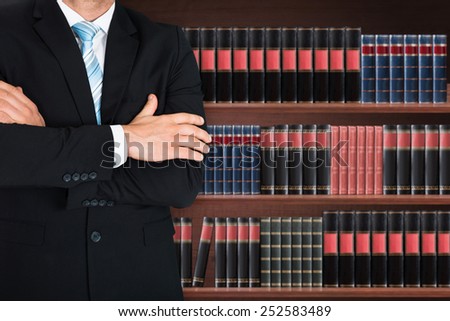 Close-up Of Male Lawyer With Arm Crossed Standing In Front Of Book Shelf