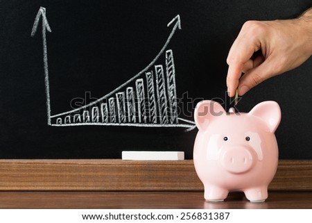 Hand Putting A Coin Into Piggy Bank In Front Of Blackboard Showing Graph