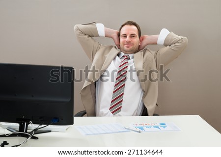 Businessman With Hands Behind Head Sitting On Office Chair At Desk