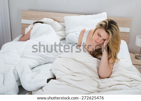 Unhappy Woman With Blanket On Bed While Man Sleeping In Bedroom
