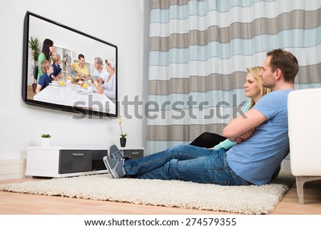 Couple Sitting On Carpet Watching Television In Living Room