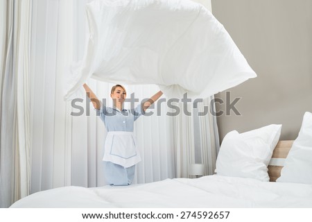 Happy Female Chambermaid Making Bed In Hotel Room