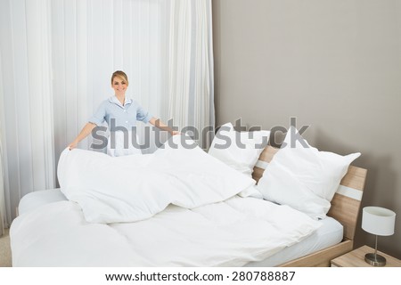 Happy Female Chambermaid Making Bed In Hotel Room
