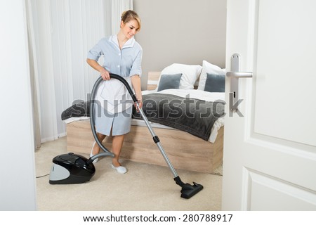 Young Maid Cleaning In Hotel Room With Vacuum Cleaner