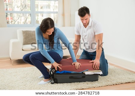 Young Couple Trying To Close Suitcase With To Much Clothes At Home