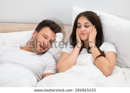 Irritated Woman Covering Her Ears While Man Snoring On Bed At Home