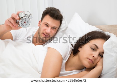 Man Lying On Bed Holding Alarm Clock To Wake Her Wife Sleeping