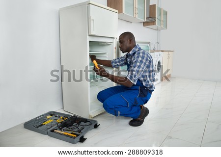Young African Repairman Repairing Refrigerator In Kitchen Room