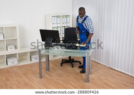 Young Male African Janitor Cleaning Desk With Cloth In Office