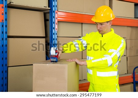 Male Worker Sealing Cardboard Box With Adhesive Tape In Warehouse
