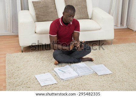 Portrait Of Young African Man Sitting On Carpet Calculating Invoices Using Calculator