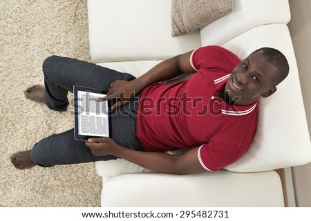 High Angle View Of Smiling Young African Man Sitting On Couch Holding Digital Tablet