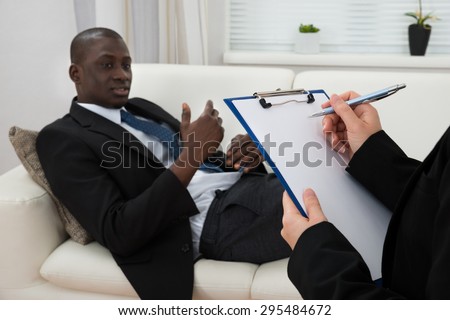 African Patient Sitting On Couch And Female Psychiatrist Writing On Clipboard
