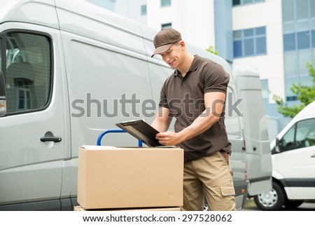 Young Happy Delivery Man With Cardboard Boxes Writing On Clipboard