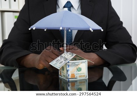 Close-up Of Businessman Hands Protecting House Of Banknote With Umbrella At Desk