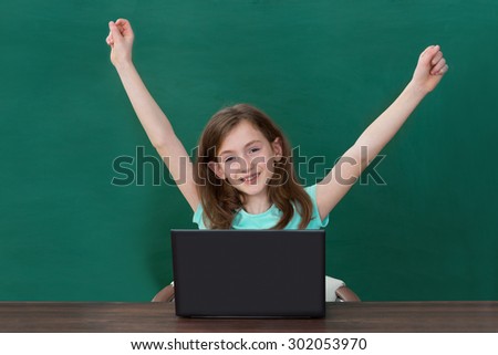 Happy Girl Raising Arms With Laptop At Desk In Classroom