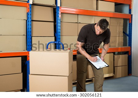 Male Worker With Cardboard Boxes Writing On Clipboard In Warehouse