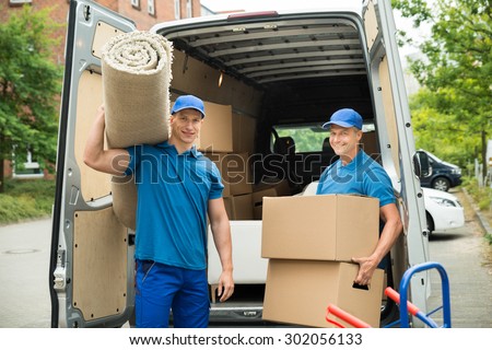 Two Male Workers Carrying Carpet And Cardboard Boxes In Front Of Van