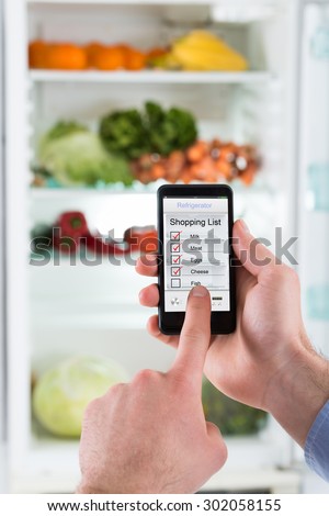 Close-up Of Person Hands Making Shopping List On Mobile Phone Display Connected To Refrigerator