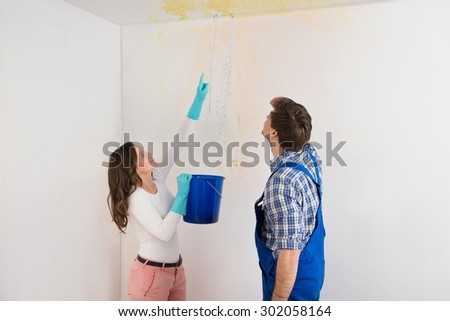Young Woman With Bucket Showing Water Leaking From Damage Ceiling To Maintenance Guy