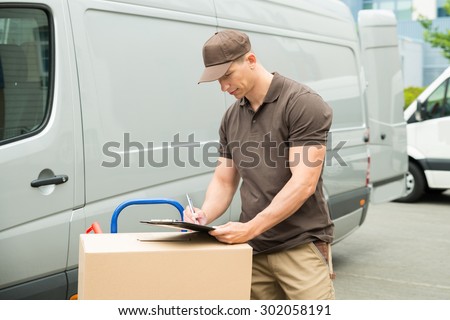 Young Happy Delivery Man With Cardboard Boxes Writing On Clipboard