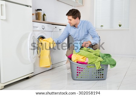 Young Man Loading Clothes Into Washing Machine In Kitchen
