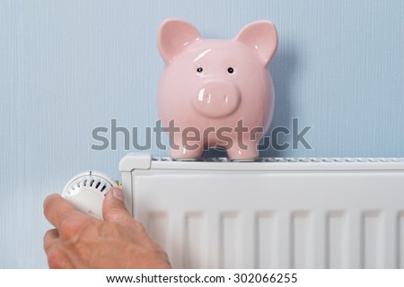 Close-up Of Man\'s Hand Adjusting Thermostat With Piggy Bank On Radiator