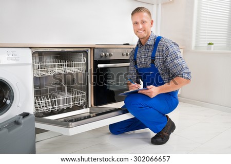 Young Handyman Looking At Dishwasher And Writing On Clipboard