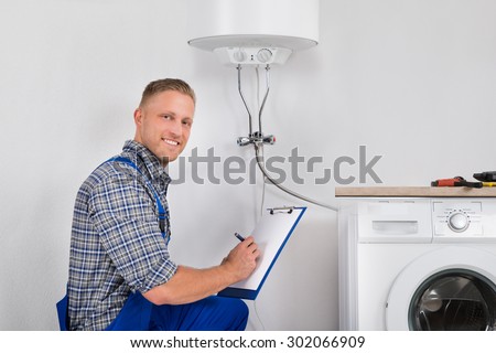Male Plumber Smiling While Writing Meter Reading Of Electric Boiler On Clipboard