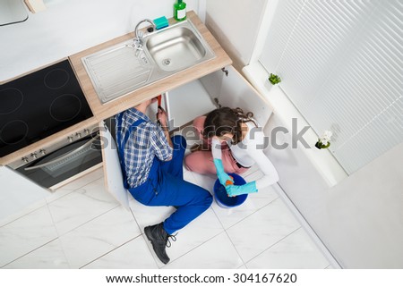 Repairman Fixing Pipe Under Sink While Woman Squeezing Wet Cloth In Kitchen