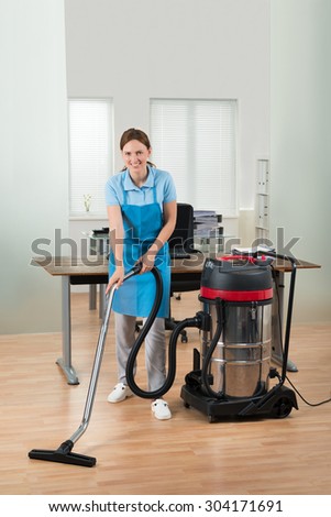 Happy Female Worker Cleaning Floor With Vacuum Cleaner In Office