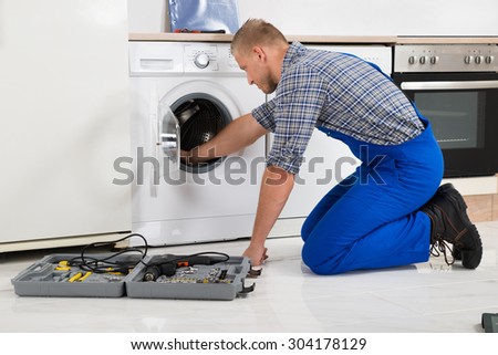 Male Worker With Toolbox Repairing Washing Machine In Kitchen