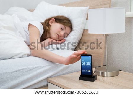 Girl Lying On Bed Snoozing Mobile Phone Alarm Clock In Bedroom